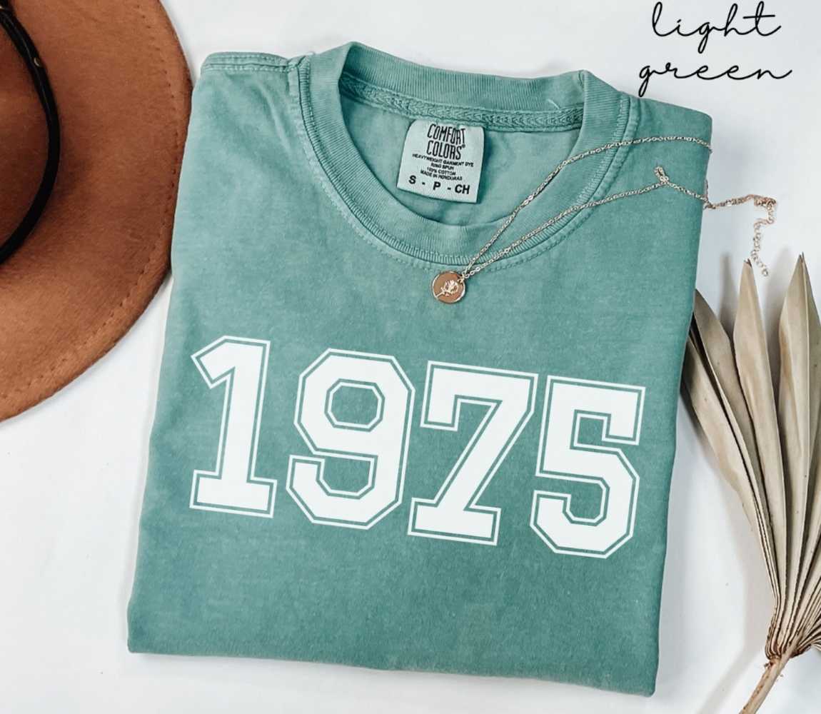 A light green t-shirt with '1975' printed on it, placed on a white surface, accompanied by a hat and dried leaves.