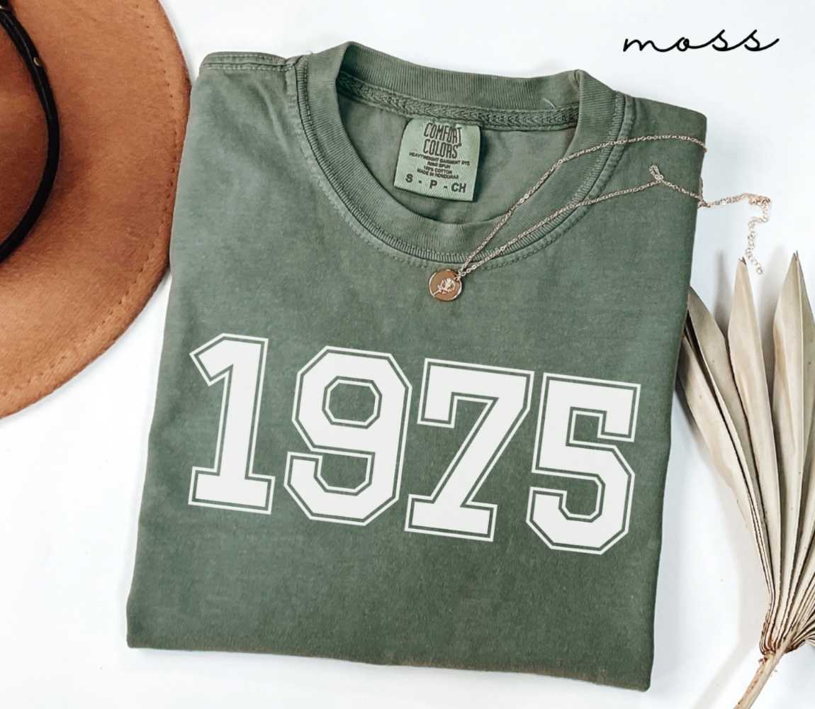 Folded green (moss) t-shirt featuring a '1975' print on a white surface alongside a hat and dried leaves.
