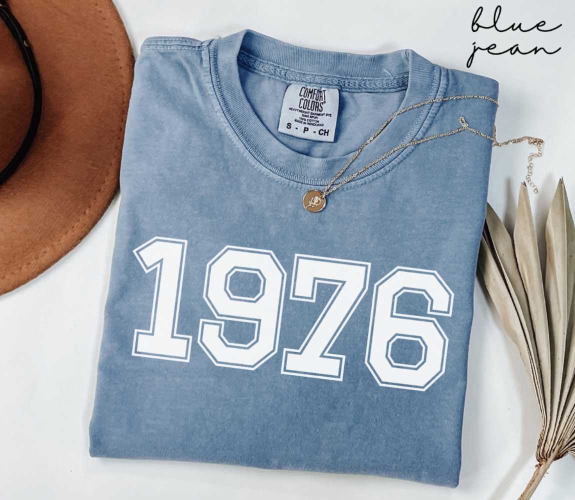 Folded blue jean t-shirt featuring '1976' print on a white surface, accompanied by a hat and dried leaves.
