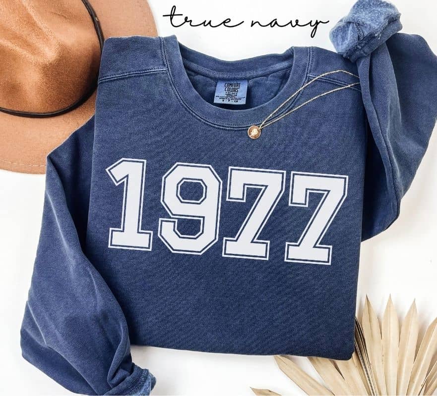 Blue (true navy) sweatshirt with '1977' printed on it, displayed with a hat and decorative elements.