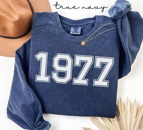 Blue (true navy) sweatshirt with '1977' printed on it, displayed with a hat and decorative elements.