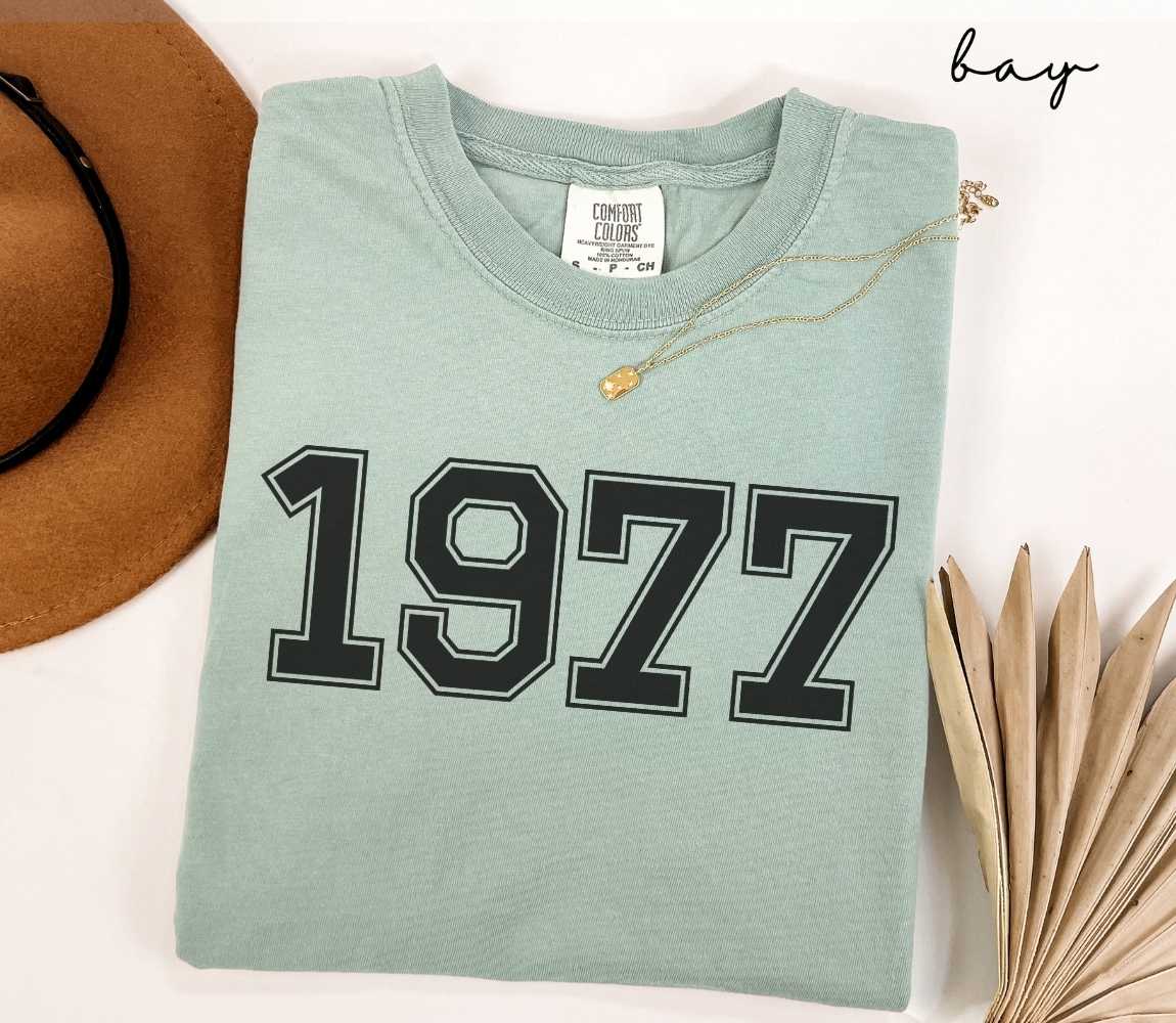 A light green (bay) t-shirt with "1977" printed on it, placed on a white surface alongside a hat and dried leaves.
