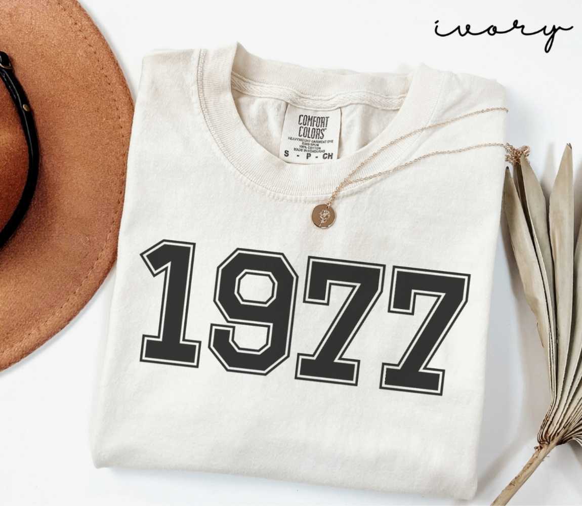 A beige (ivory) t-shirt with '1977' print, placed next to a brown hat and a dried plant on a light background.
