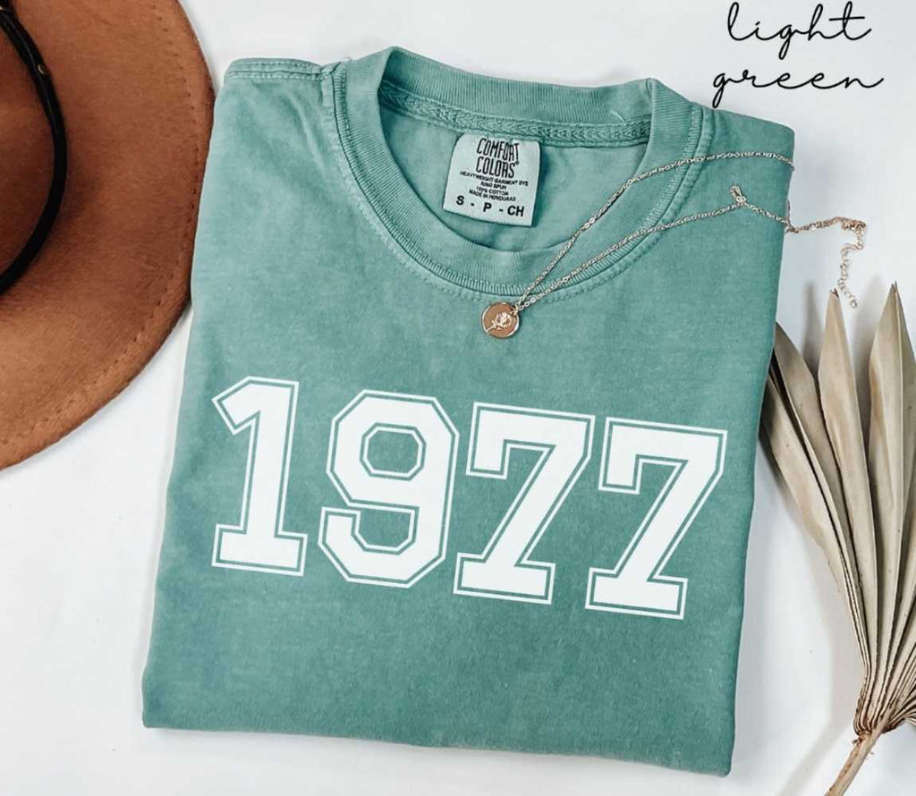 A light green t-shirt with '1977' printed on it, placed on a white surface, accompanied by a hat and dried leaves.
