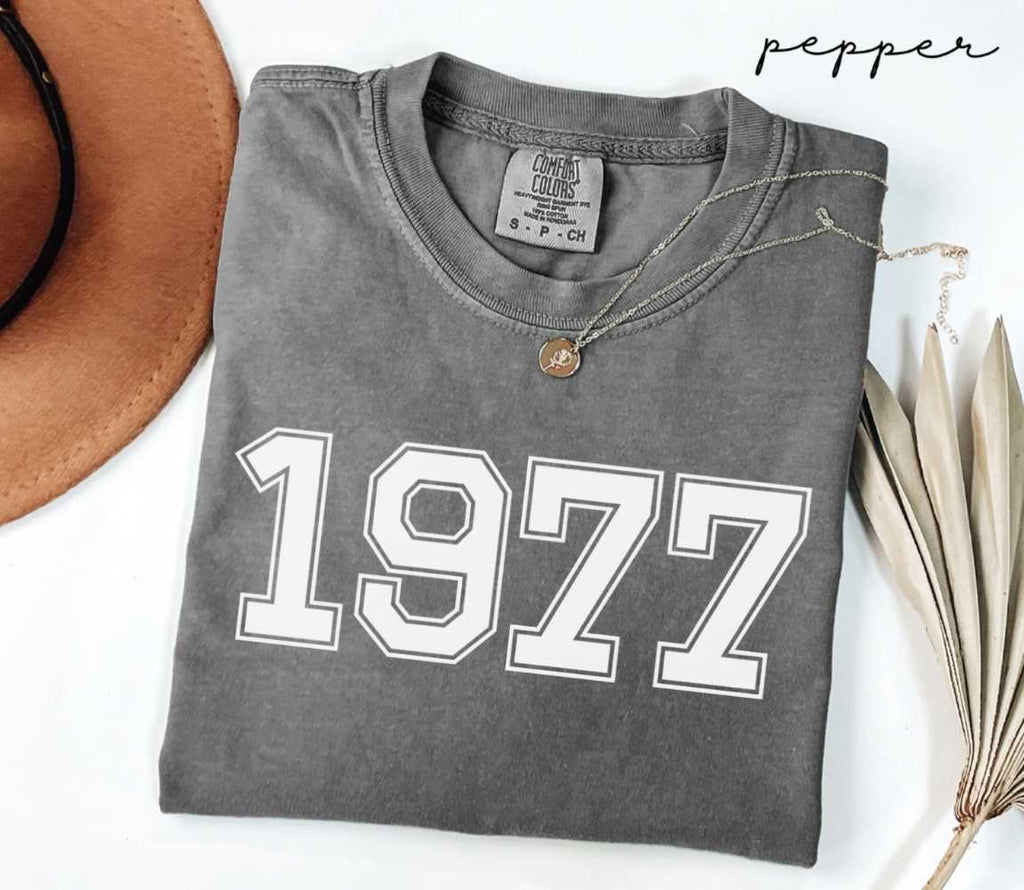Folded gray (pepper) t-shirt with '1977' print on a white surface with a hat and dried leaves.
