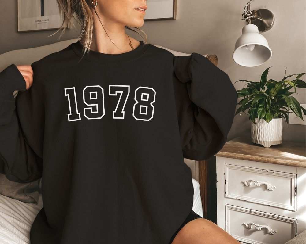 A person wearing a black sweatshirt featuring '1978' printed on it is in a bedroom.