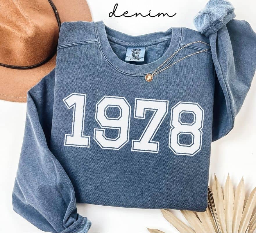 Blue (denim) sweatshirt with '1978' printed on it, worn by a person, with a plain background.