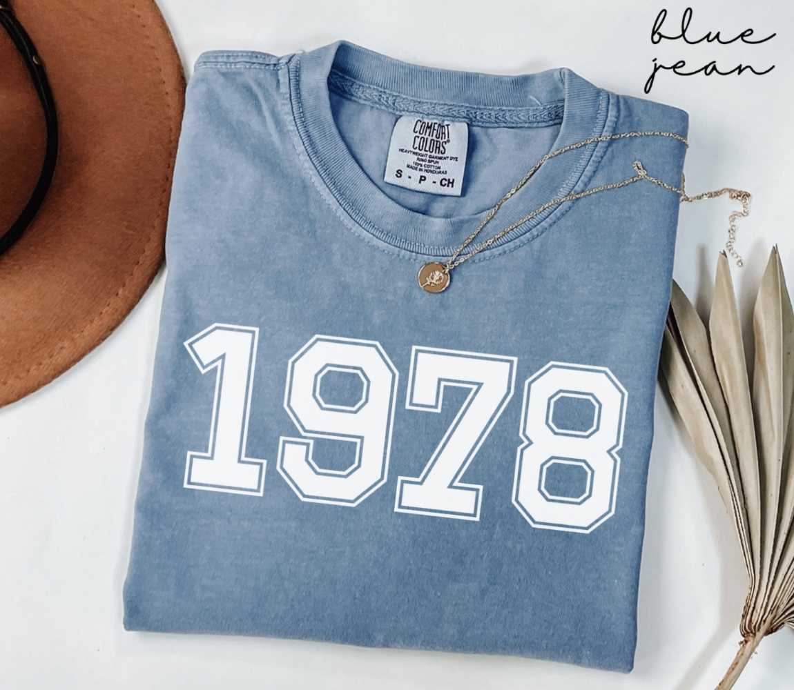 Folded blue jean t-shirt featuring '1978' print on a white surface, accompanied by a hat and dried leaves.

