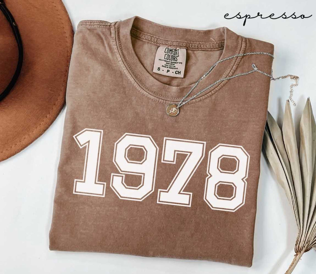 Folded brown (espresso) t-shirt with '1978' print on a white surface, accompanied by a hat and dried leaves.

