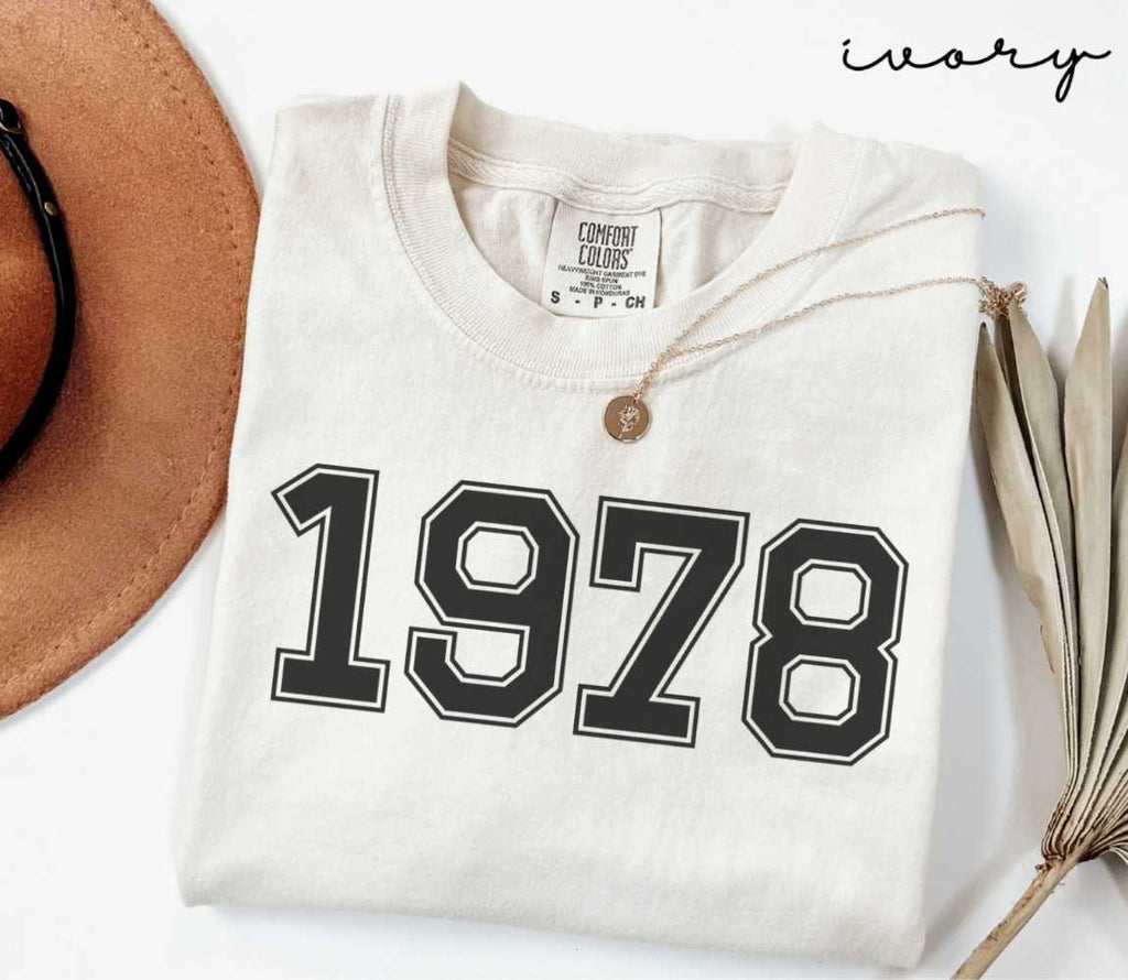 A beige (ivory) t-shirt with '1978' print, placed next to a brown hat and a dried plant on a light background.
