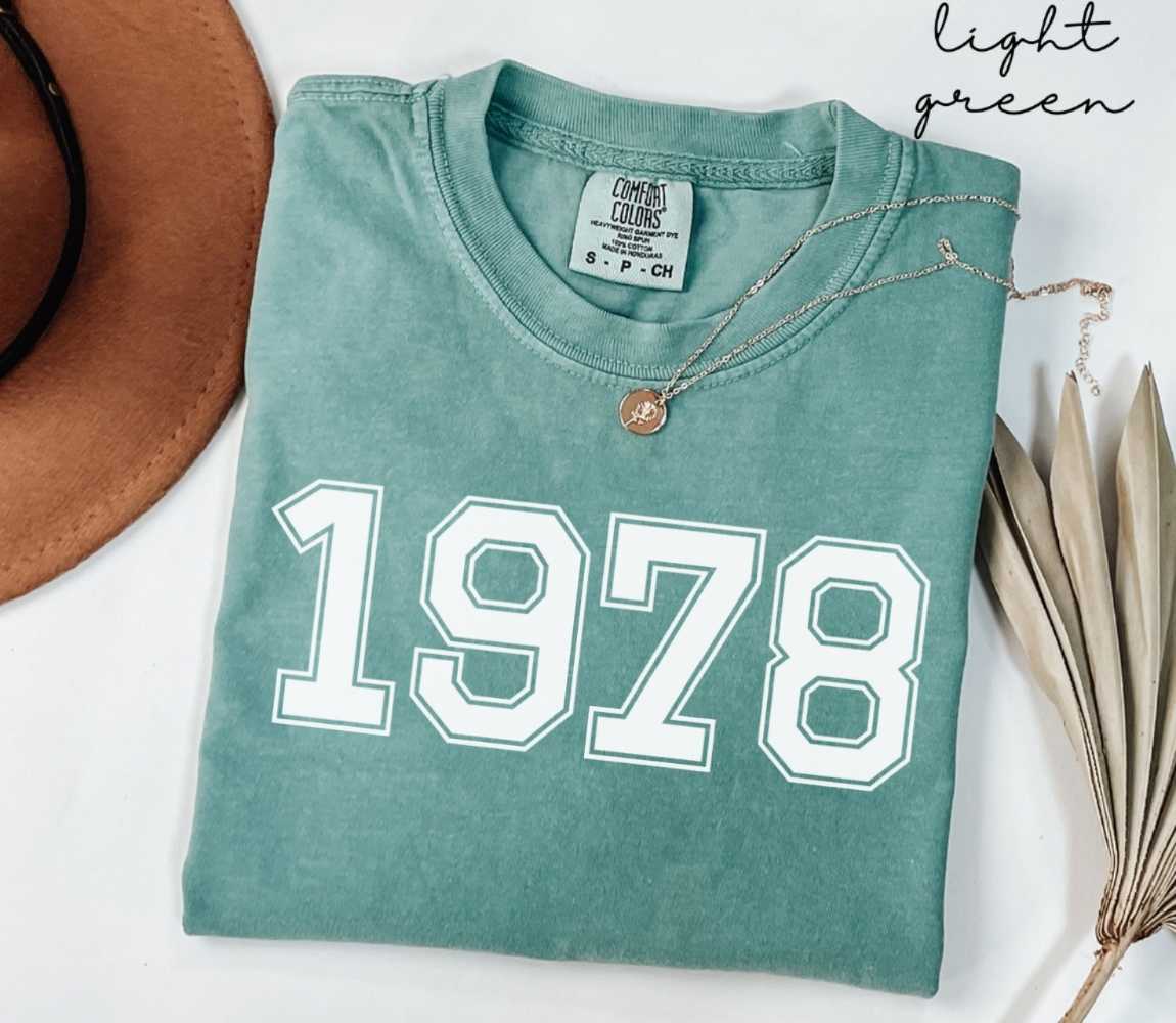 A light green t-shirt with '1978' printed on it, placed on a white surface, accompanied by a hat and dried leaves.
