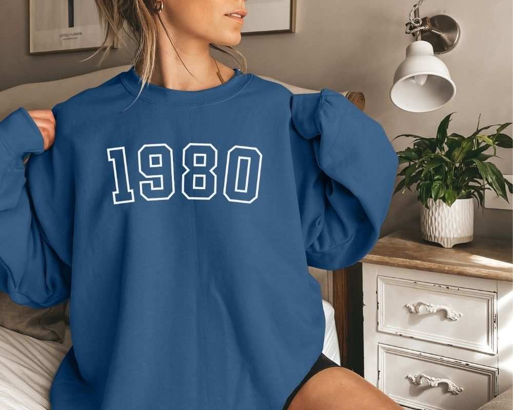 A person wearing an indigo blue sweatshirt with '1980' printed on it in a bedroom setting.