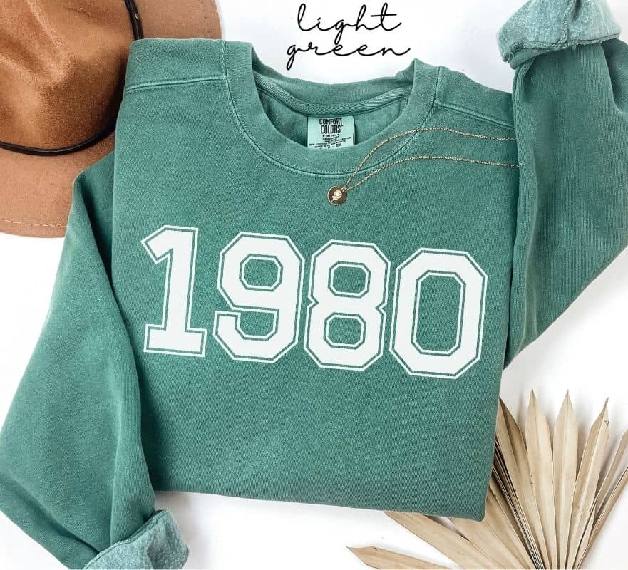 Light green sweatshirt with '1980' printed on it, displayed on a white background.