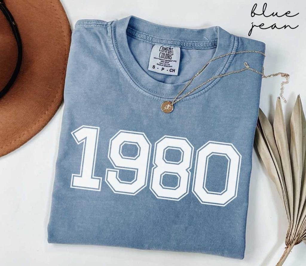 Folded blue jean t-shirt featuring '1980' print on a white surface, accompanied by a hat and dried leaves.
