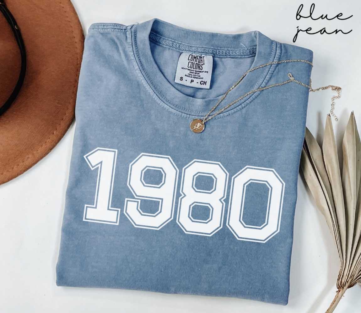 Folded blue jean t-shirt featuring '1980' print on a white surface, accompanied by a hat and dried leaves.
