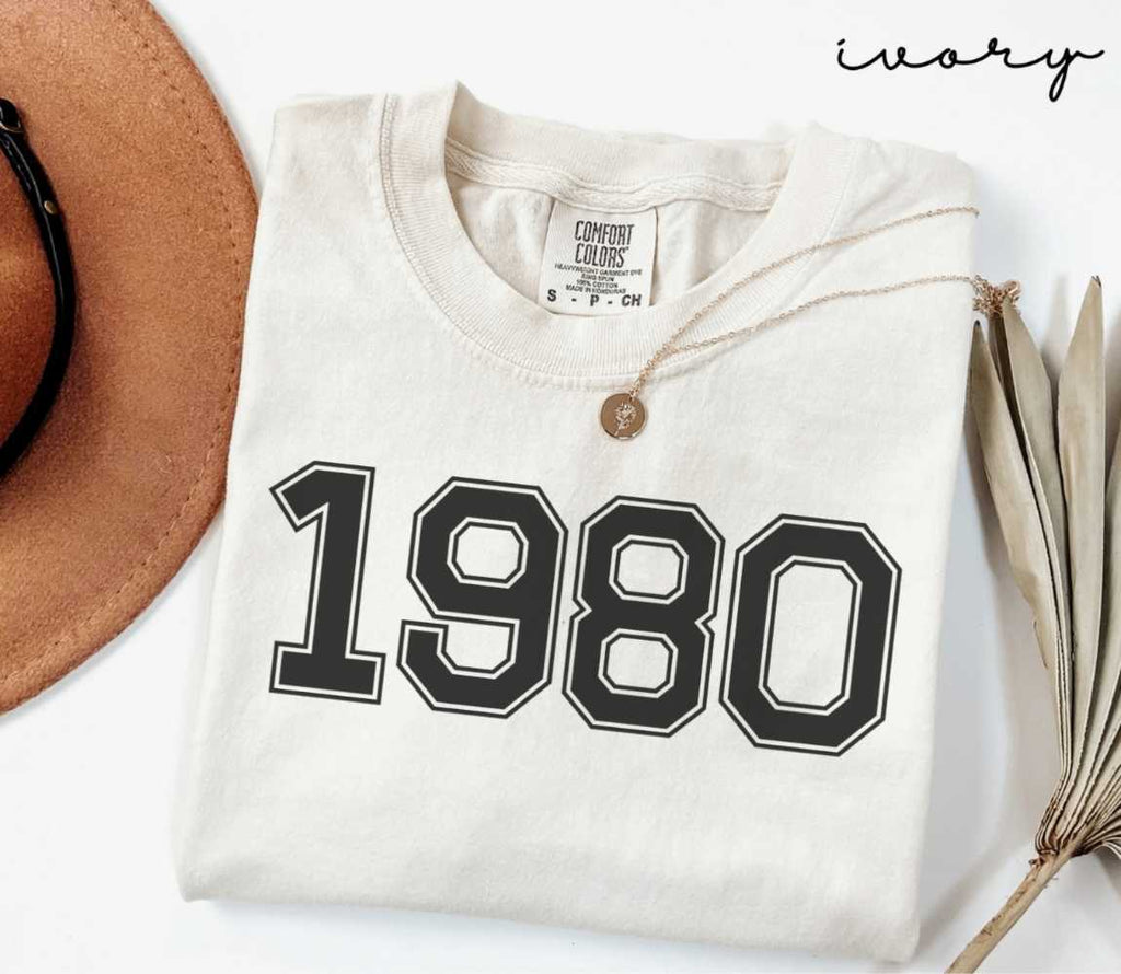 A beige (ivory) t-shirt with '1980' print, placed next to a brown hat and a dried plant on a light background.
