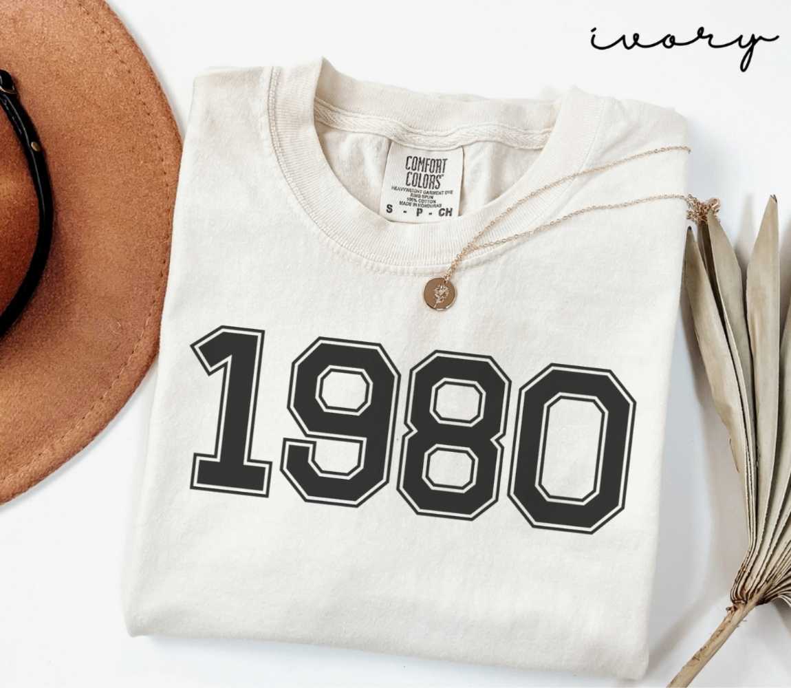 A beige (ivory) t-shirt with '1980' print, placed next to a brown hat and a dried plant on a light background.

