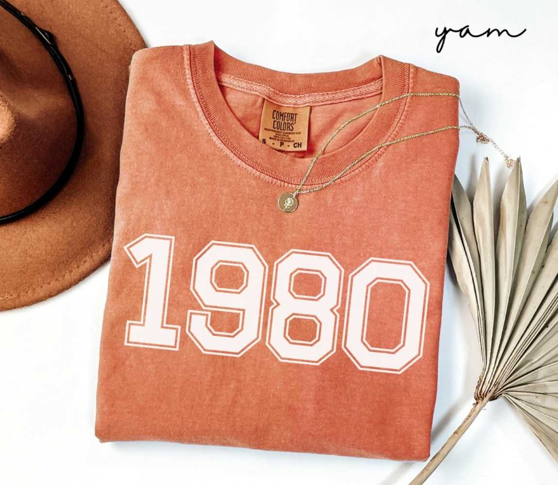Folded orange (yam) t-shirt with a '1980' print, a brown hat, and a dried plant on a white background.
