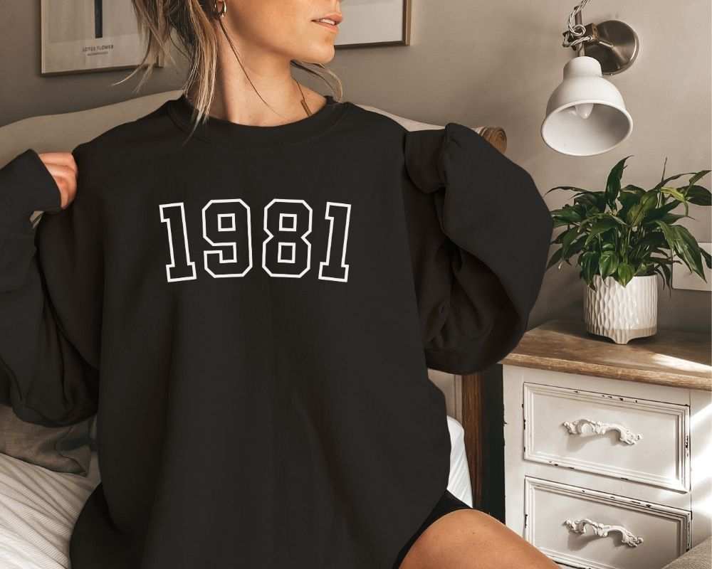 A person is sitting in a room, wearing a black sweatshirt that has '1981' printed on it.