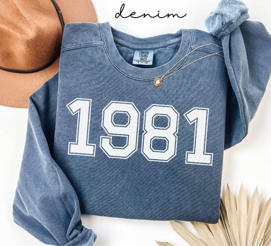 Blue (denim) sweatshirt with '1981' printed on it, worn by a person, with a plain background.