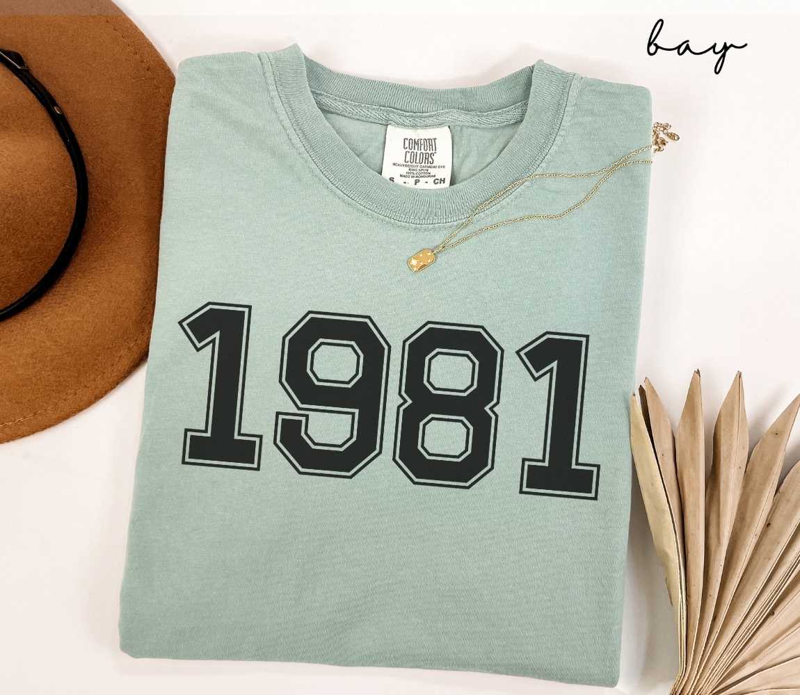 A light green (bay) t-shirt with "1981" printed on it, placed on a white surface alongside a hat and dried leaves.
