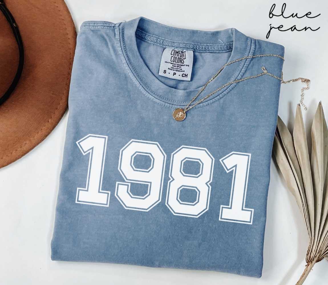 Folded blue jean t-shirt featuring '1981' print on a white surface, accompanied by a hat and dried leaves.
