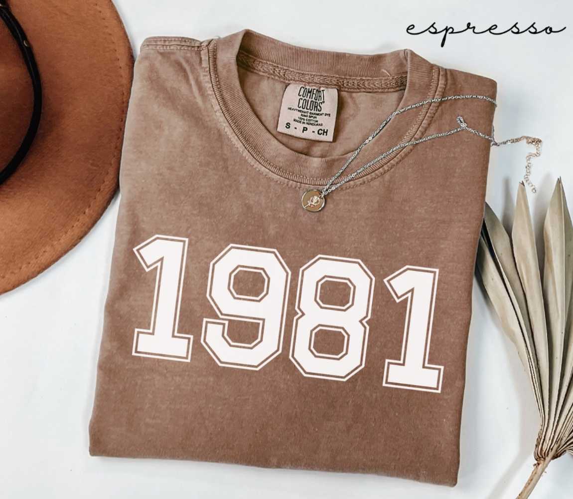 Folded brown (espresso) t-shirt with '1981' print on a white surface, accompanied by a hat and dried leaves.
