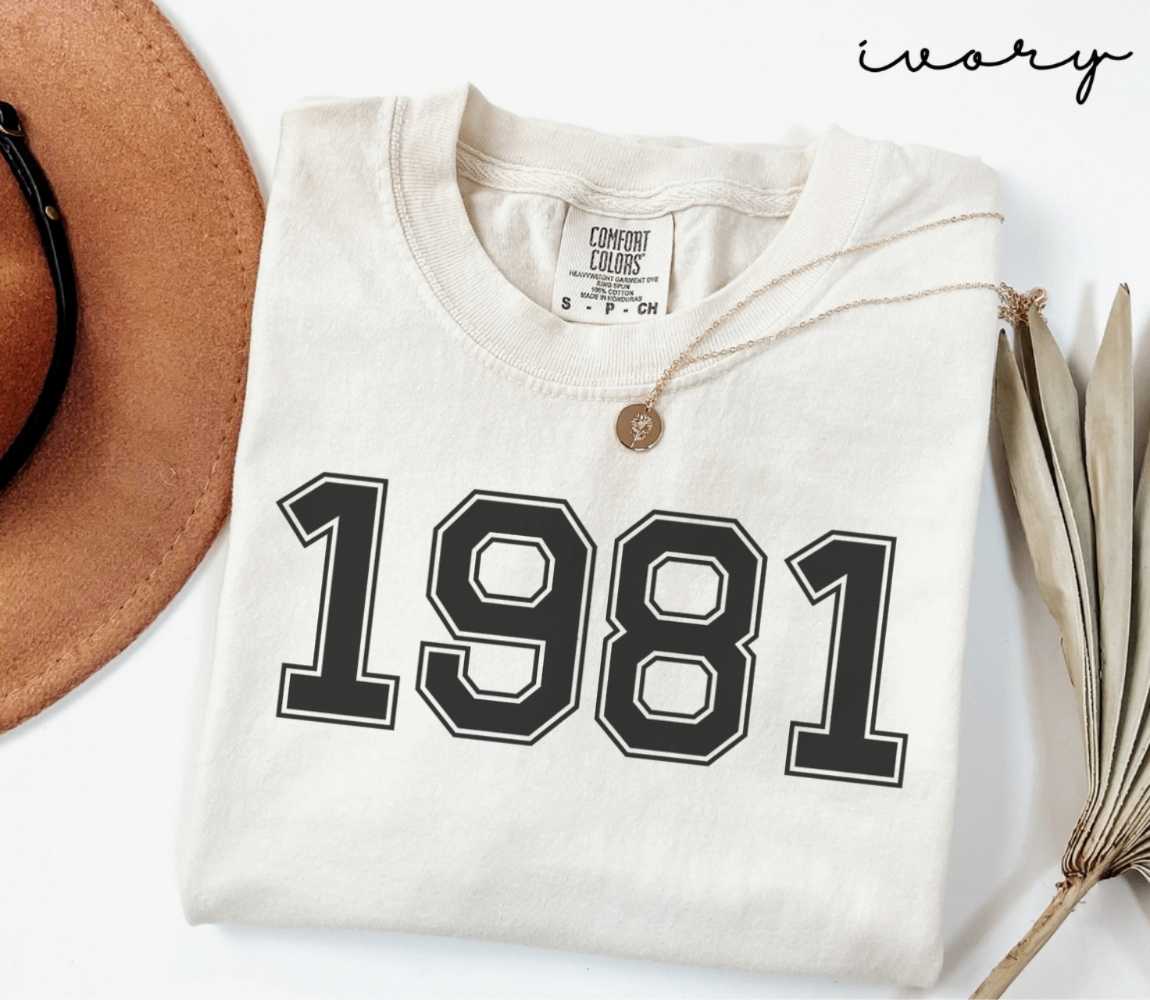 A beige (ivory) t-shirt with '1981' print, placed next to a brown hat and a dried plant on a light background.
