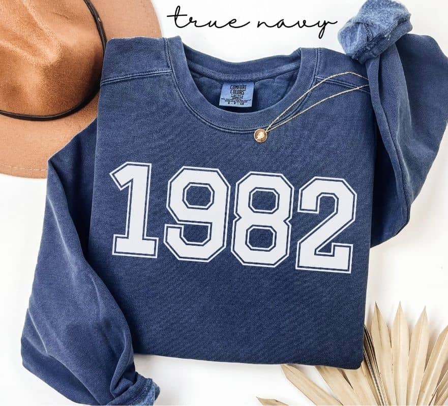 Blue (true navy) sweatshirt with '1982' printed on it, displayed with a hat and decorative elements.