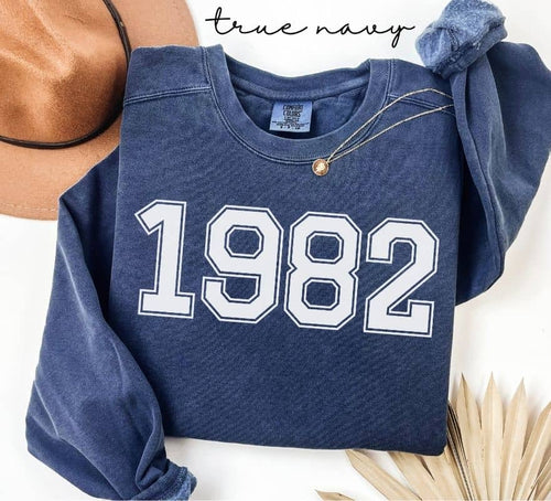 Blue (true navy) sweatshirt with '1982' printed on it, displayed with a hat and decorative elements.