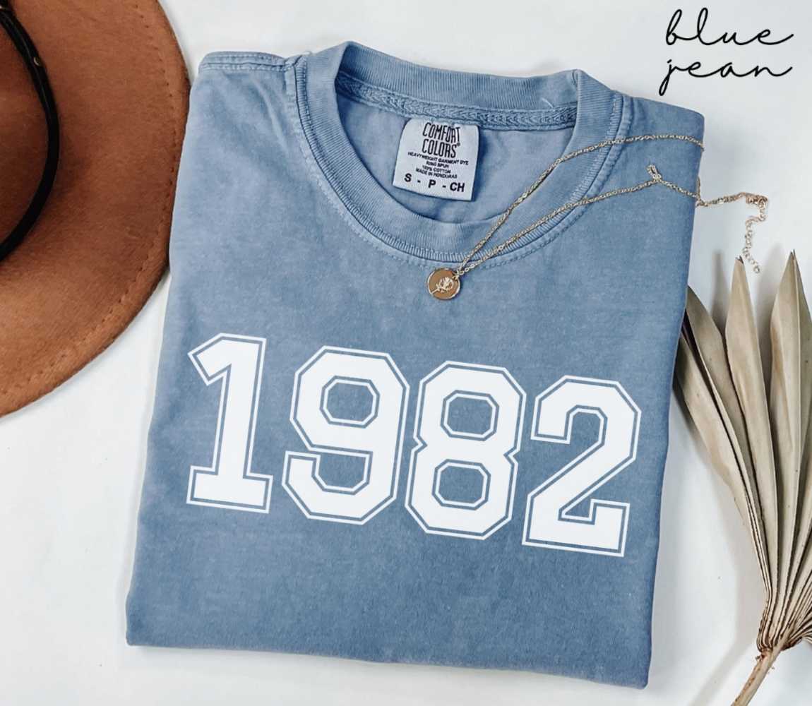 Folded blue jean t-shirt featuring '1982' print on a white surface, accompanied by a hat and dried leaves.
