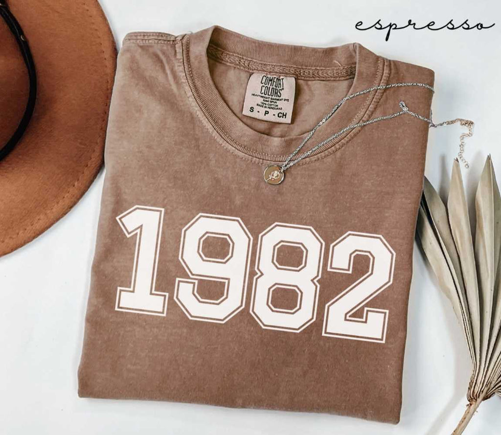 Folded brown (espresso) t-shirt with '1982' print on a white surface, accompanied by a hat and dried leaves.

