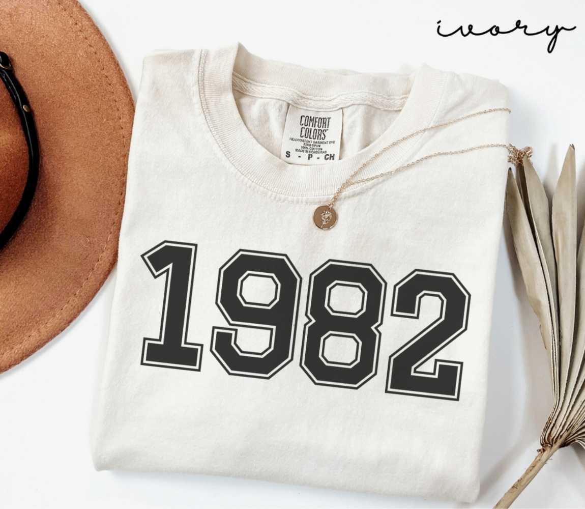 A beige (ivory) t-shirt with '1982' print, placed next to a brown hat and a dried plant on a light background.

