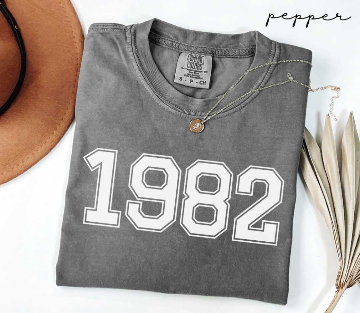 Folded gray (pepper) t-shirt with '1982' print on a white surface with a hat and dried leaves.
