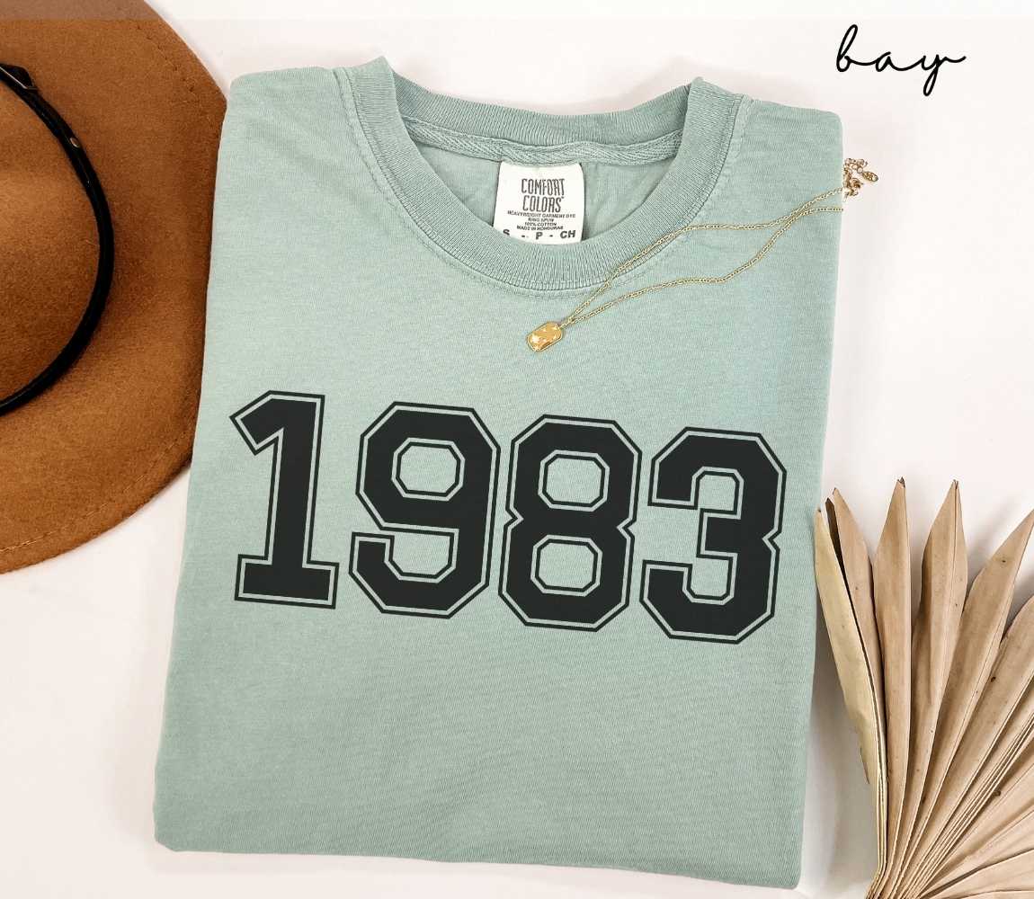 A light green (bay) t-shirt with "1983" printed on it, placed on a white surface alongside a hat and dried leaves.
