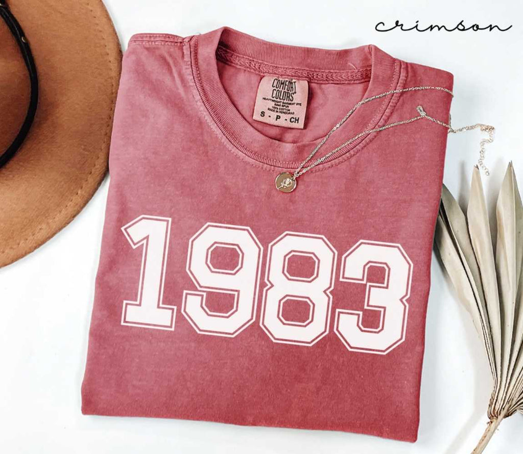 A red (Crimson) folded t-shirt with '1983' printed on it, next to a hat, and dried leaves on a white background.

