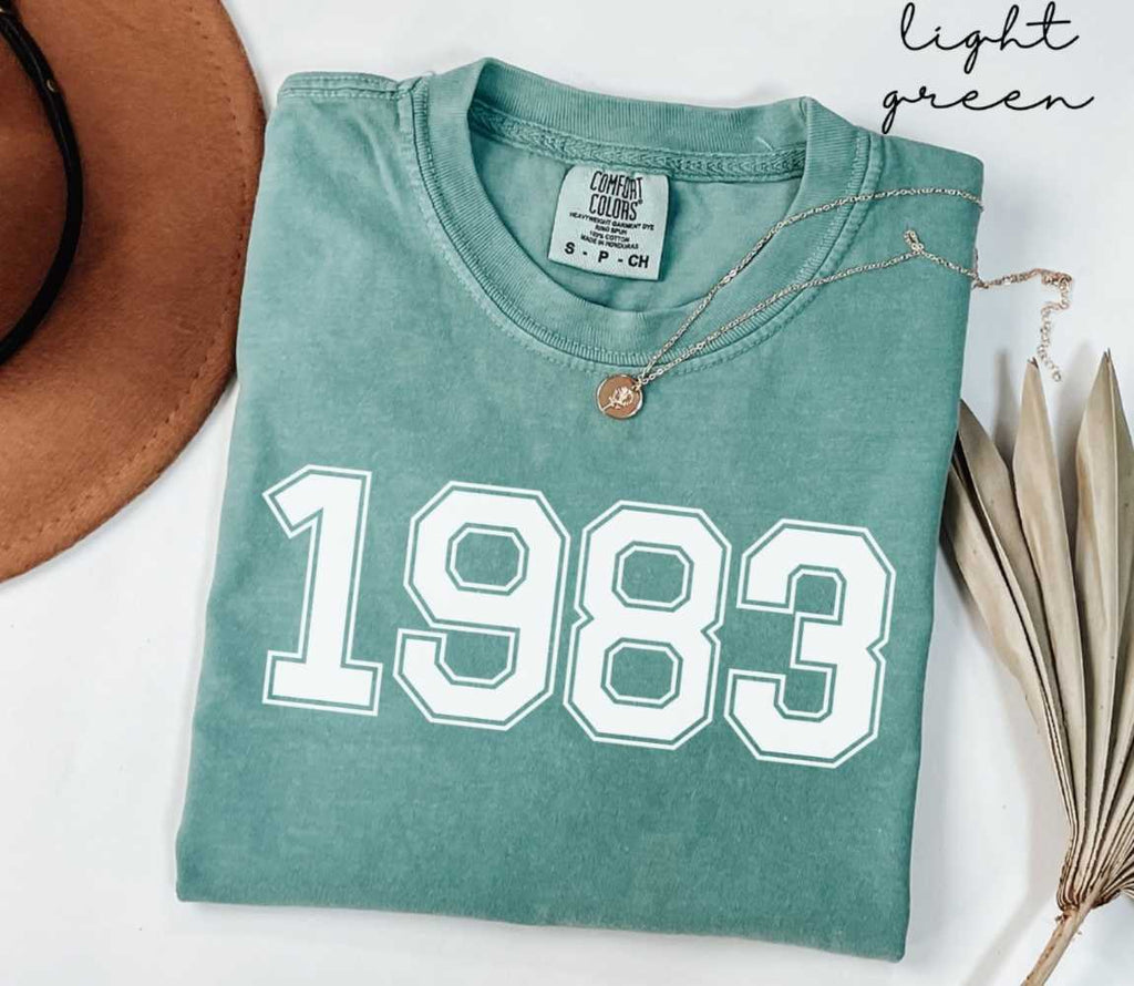 A light green t-shirt with '1983' printed on it, placed on a white surface, accompanied by a hat and dried leaves.
