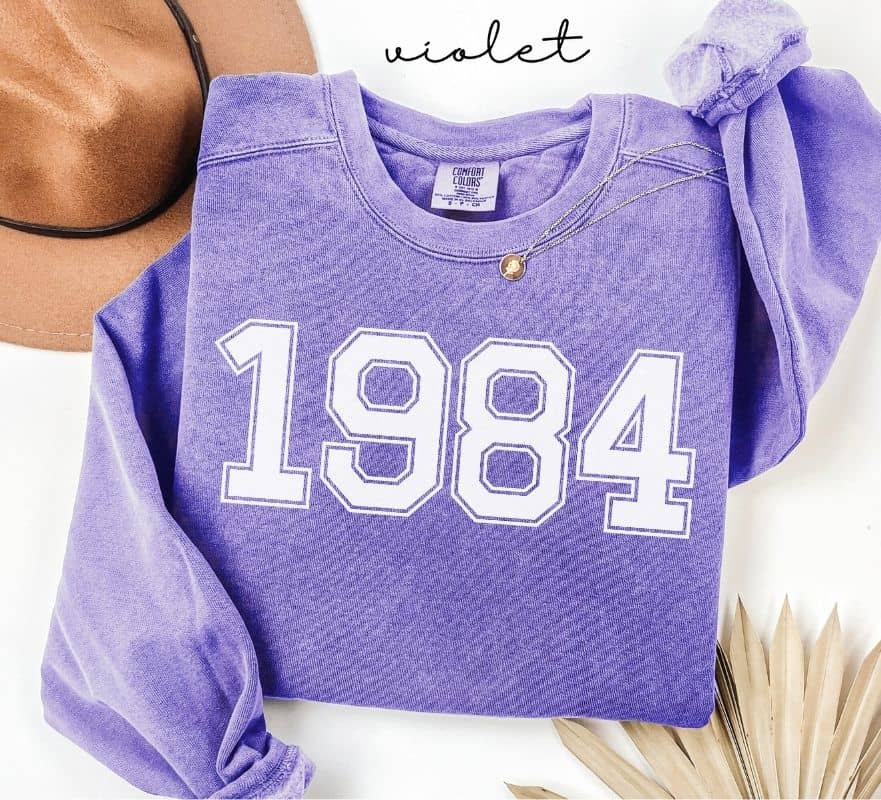 Purple (violet) sweatshirt with '1984' printed on it, worn by a person, with a plain background.