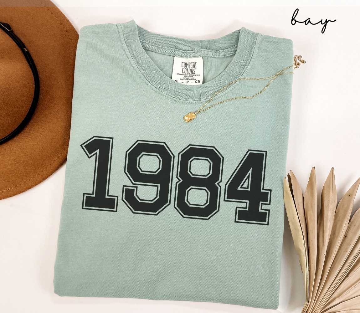 A light green (bay) t-shirt with "1984" printed on it, placed on a white surface alongside a hat and dried leaves.

