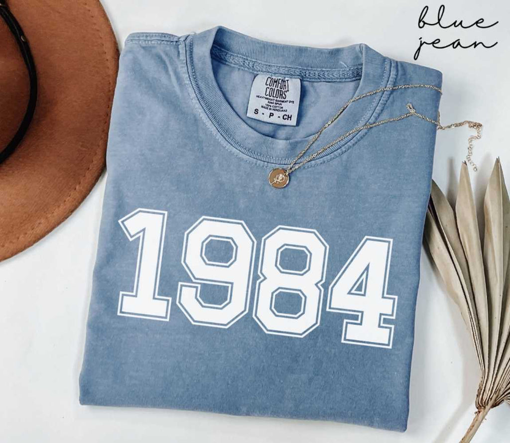 Folded blue jean t-shirt featuring '1984' print on a white surface, accompanied by a hat and dried leaves.
