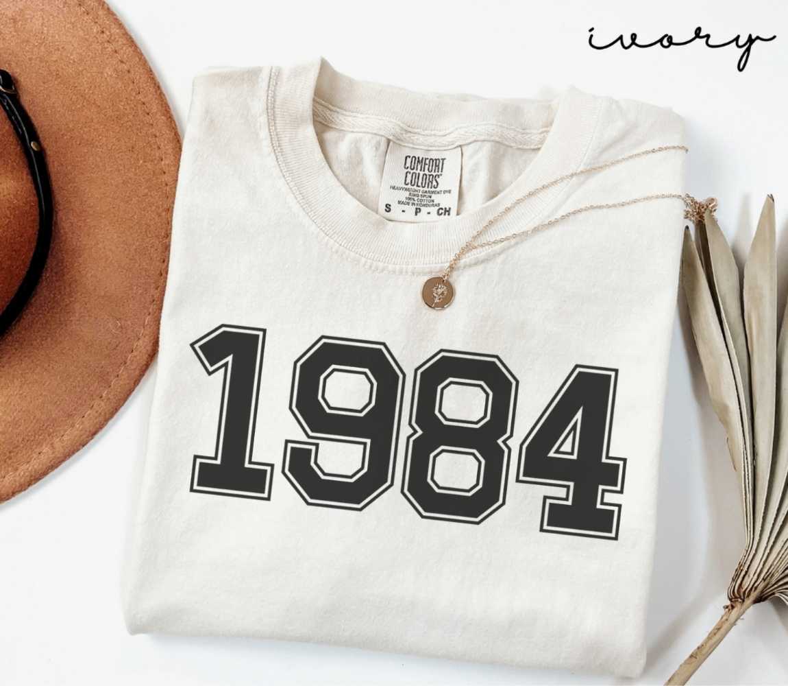 A beige (ivory) t-shirt with '1984' print, placed next to a brown hat and a dried plant on a light background.
