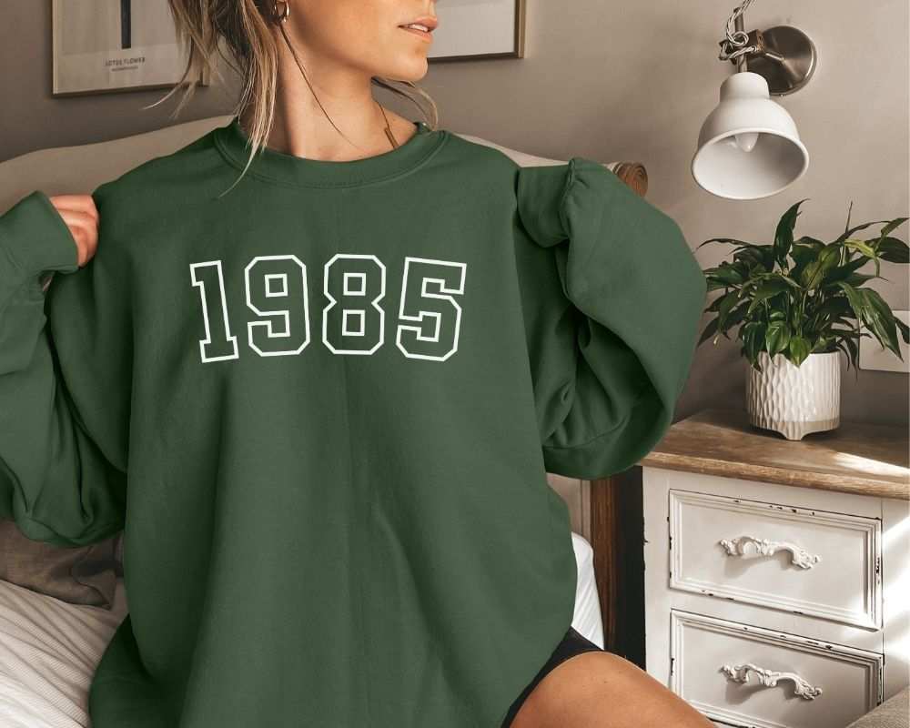 A person is sitting in a bedroom, wearing a forest green sweatshirt that has '1985' printed on it.