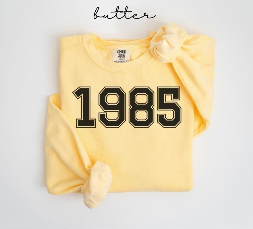 Yellow (butter) sweatshirt with '1985' printed on it, folded and displayed against a white background.