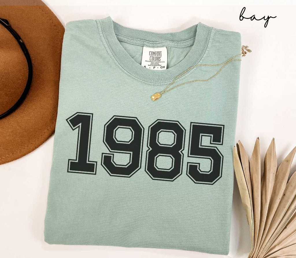 A light green (bay) t-shirt with "1985" printed on it, placed on a white surface alongside a hat and dried leaves.
