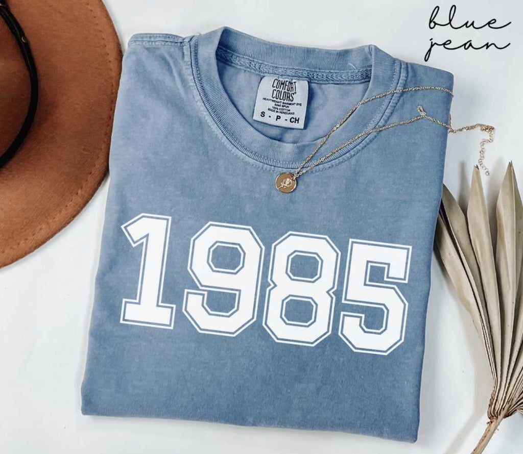 Folded blue jean t-shirt featuring '1985' print on a white surface, accompanied by a hat and dried leaves.
