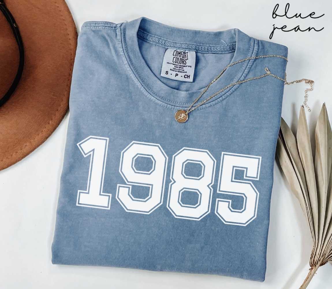 Folded blue jean t-shirt featuring '1985' print on a white surface, accompanied by a hat and dried leaves.
