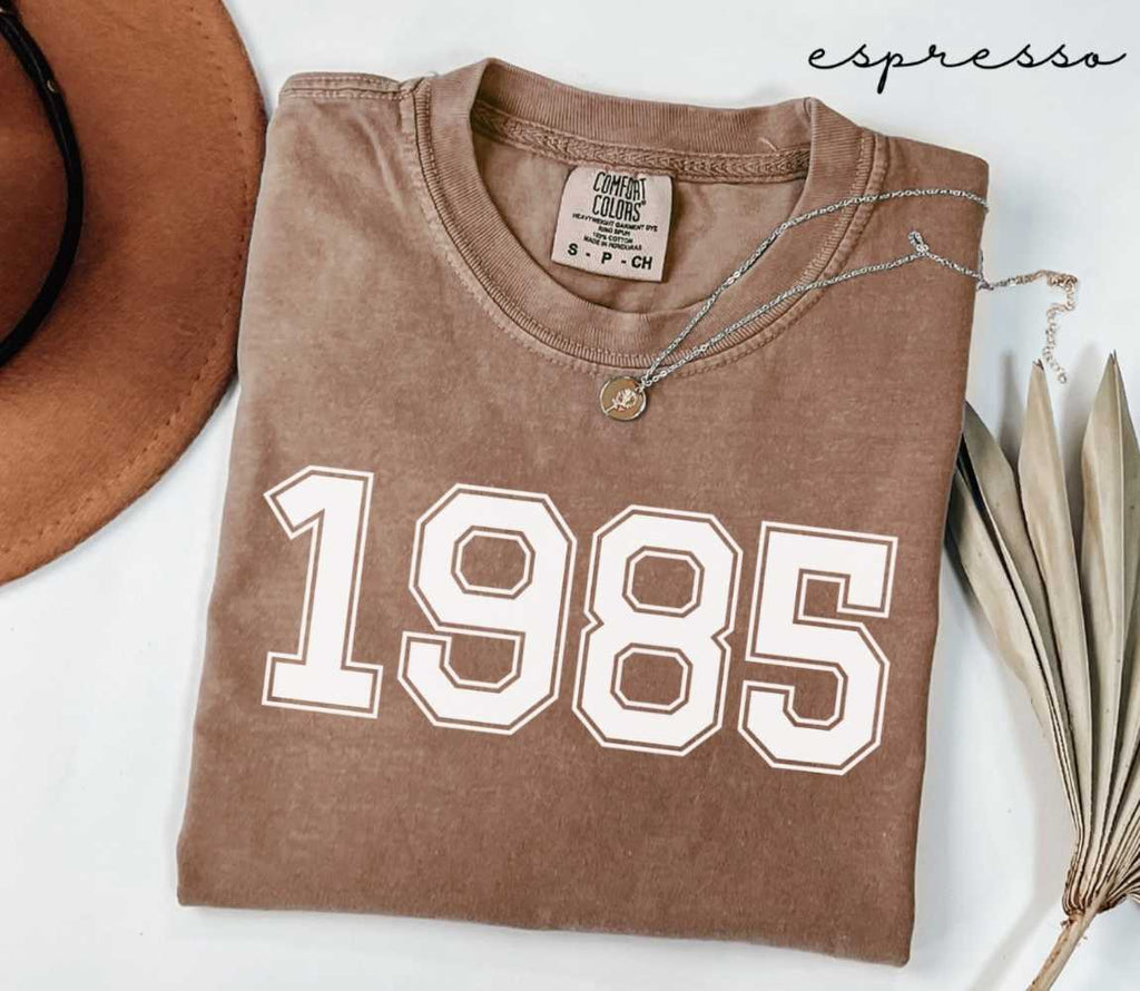Folded brown (espresso) t-shirt with '1985' print on a white surface, accompanied by a hat and dried leaves.

