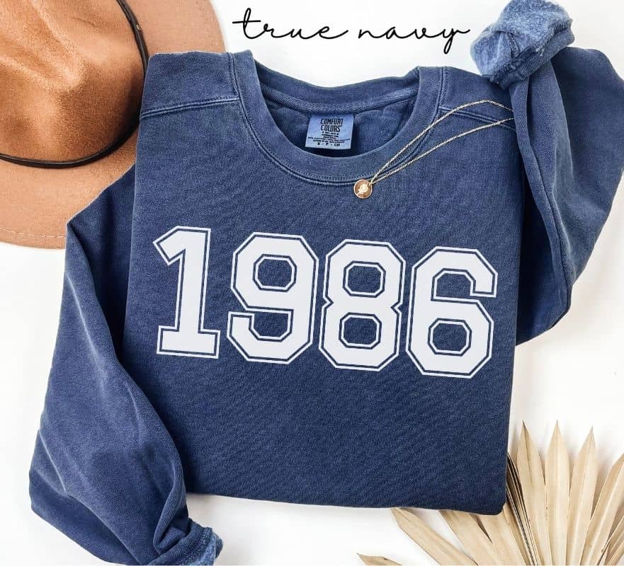 Blue (true navy) sweatshirt with '1986' printed on it, displayed with a hat and decorative elements.