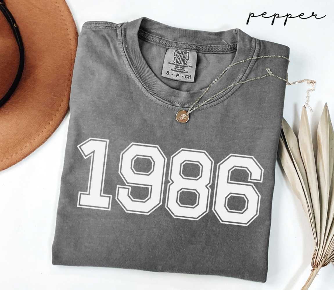 Folded gray (pepper) t-shirt with '1986' print on a white surface with a hat and dried leaves.
