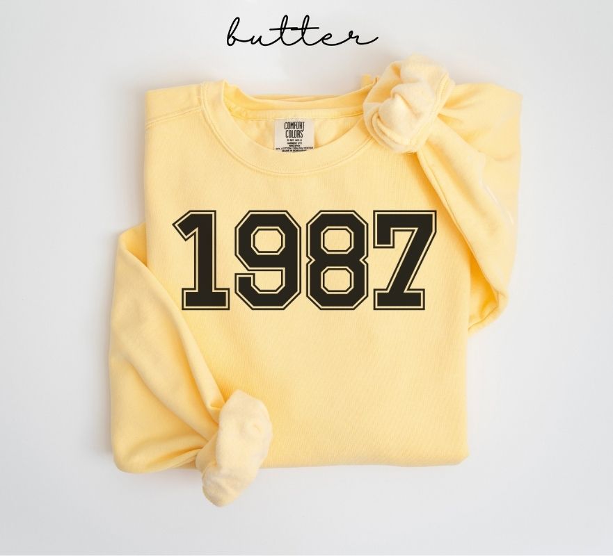 Yellow (butter) sweatshirt with '1987' printed on it, folded and displayed against a white background.
